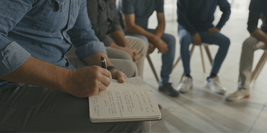 A group of individuals seated in a circle, with a man actively writing notes on a notepad during a discussion.
