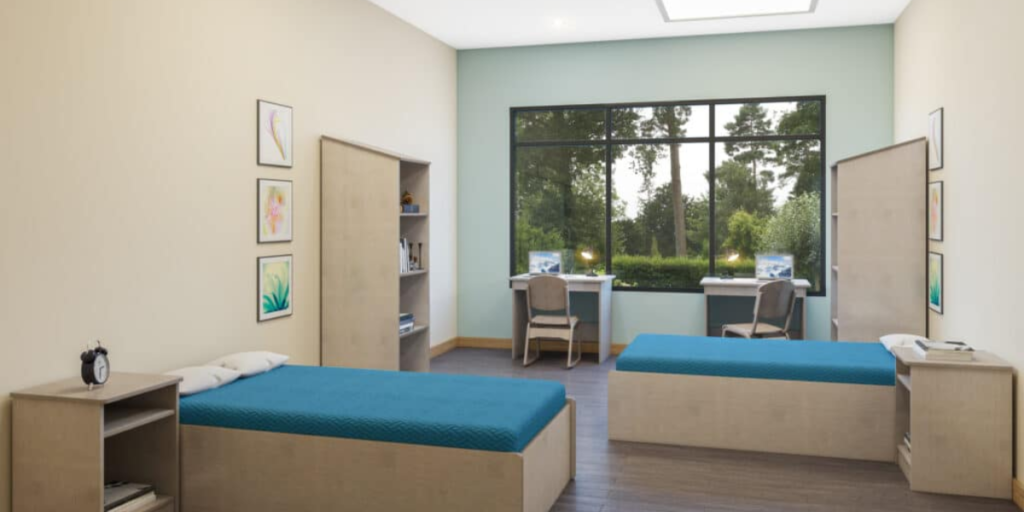 A room featuring two beds and a desk, designed for comfort and functionality in residential units and patient care settings.