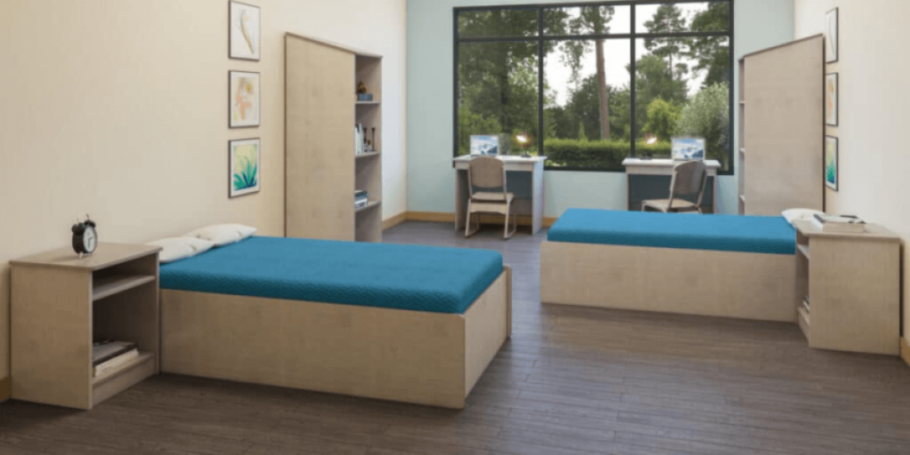 A room featuring two beds and a desk, designed for group living spaces with reliable, ready-to-ship furniture.