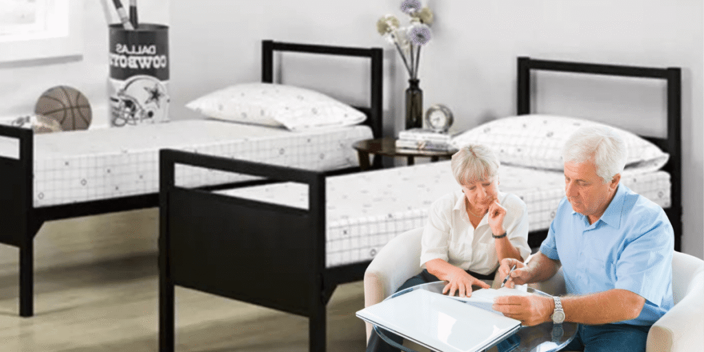 An older couple sits on a bed, using a laptop, possibly discussing furniture costs and tariffs affecting their budget.